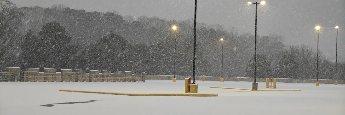 An empty parking deck under heavy falling snow at dusk — unbroken white ground, a line of lamp posts glowing sodium-yellow against grey sky and dark winter trees.